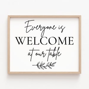 May include: A framed print with the words "Everyone is WELCOME at our table" in black script and block letters. A decorative sprig of leaves is below the text. The frame is a light wood color.