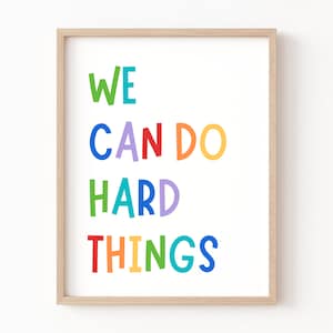 以下が含まれることがあります： 虹色の文字で「WE CAN DO HARD THINGS」と書かれた額装されたプリント。文字は遊び心のある手書き風です。