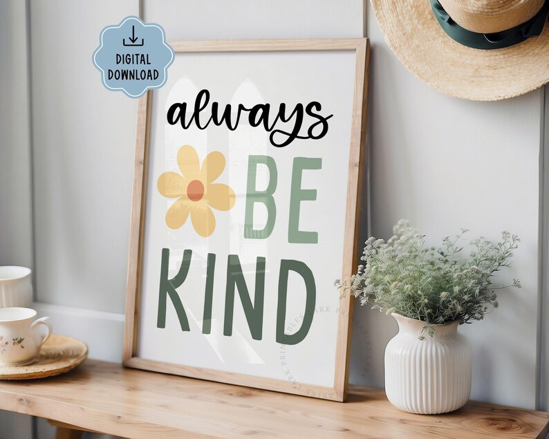 May include: A framed print with the words "always BE KIND" in black and green. A yellow flower graphic is between "always" and "BE". The print is in a light wood frame and is on a wooden surface.