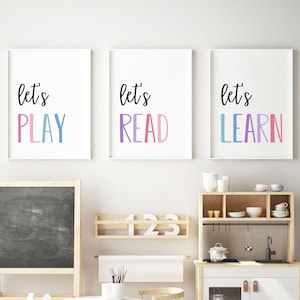 Puede incluir: Tres impresiones enmarcadas con las frases "let's PLAY", "let's READ" y "let's LEARN" en escritura negra y letras de bloque coloridas. Las impresiones se muestran encima de un área de juegos para niños con una pizarra, una cocina de juguete y una estantería de madera.
