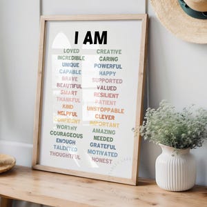 May include: A framed print with the words "I AM" in large black letters at the top. The print features a list of positive affirmations in colorful text, including words like "loved", "creative", "incredible", "caring", "unique", "powerful", "capable", "happy", "brave", "supported", "beautiful", "valued", "smart", "resilient", "thankful", "patient", "kind", "unstoppable", "helpful", "clever", "confident", "important", "worthy", "amazing", "courageous", "needed", "enough", "grateful", "talented", "motivated", "thoughtful", "honest".