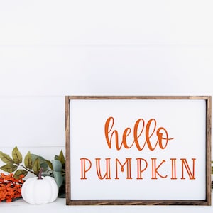 May include: A white wooden sign with a brown frame, featuring the text "hello pumpkin" in orange lettering. The sign is sitting on a white surface with a white pumpkin and orange flowers.