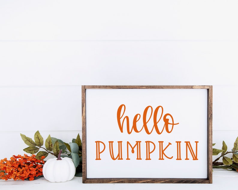 May include: A white wooden sign with a brown frame, featuring the text "hello pumpkin" in orange lettering. The sign is sitting on a white surface with a white pumpkin and orange flowers.