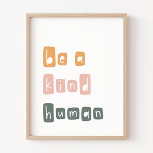 Puede incluir: Impresi&oacute;n enmarcada con la frase "Be a kind human" en una fuente l&uacute;dica y en bloques. El texto est&aacute; dispuesto en tres l&iacute;neas, con las palabras en naranja, rosa y verde. El marco es de color madera clara.