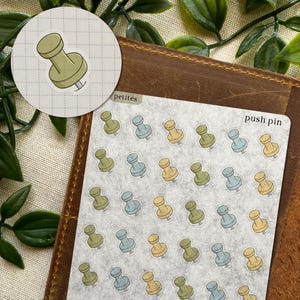 May include: A sheet of petite push pin stickers in various colours, including green, blue, and yellow. The stickers are on a textured background, with the words "push pin" printed on the sheet. A single push pin sticker is shown on a circular background.