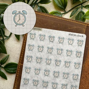 May include: A sheet of petite alarm clock stickers in a light blue and cream color scheme. The stickers feature a classic alarm clock design. The words "alarm clock" are printed on the sheet. The stickers are on a brown leather surface.