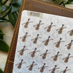 May include: Sheet of stickers featuring small brown violins with bows. The word "PETITES" is printed in a rectangular label at the top. The stickers are on a white background, and the sheet is placed on a brown leather-like surface.