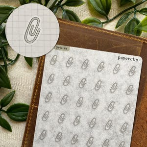 May include: A sheet of paperclip stickers with a light gray background. The word "paperclip" is printed on the top right. A close-up of a single paperclip sticker is in a circle on the top left. The stickers are on a brown leather surface.