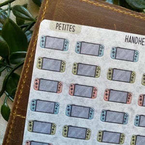 May include: A sheet of stickers featuring small illustrations of handheld gaming consoles. The stickers have a gray screen and colorful controllers in blue, pink, and green. The word "PETITES" is printed at the top of the sheet.