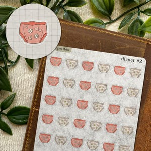 May include: A sticker sheet labeled petites diaper #2 featuring multiple small, individual stickers of diapers in two color variations: solid pink with white flower and heart patterns, and light beige with small flower and heart designs.