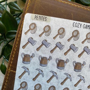 May include: A sheet of stickers with a cozy camping theme. The stickers feature illustrations of fishing nets, axes, watering cans, and pickaxes. The word "PETITES" is printed on a small label. The words "COZY CAMP" are also visible.