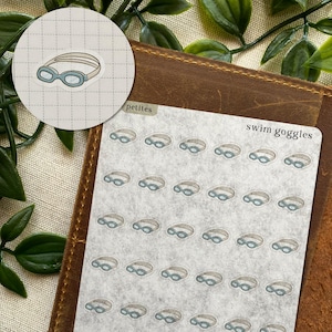 May include: A sticker sheet featuring multiple small illustrations of light blue and beige swim goggles arranged in a grid pattern. The text "petites" and "swim goggles" is printed in black on the top portion of the translucent sticker sheet.