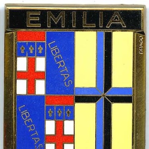 Puede incluir: Una insignia de metal dorado con forma de escudo. El escudo está dividido en cuatro secciones, cada una con un color y diseño diferente. La sección superior es negra con la palabra "EMILIA" en letras doradas. La sección inferior es amarilla. Las secciones izquierda y derecha son azules con una cruz blanca y la palabra "LIBERTAS" en letras blancas. La insignia tiene la palabra "CAL323" en letras doradas en la parte inferior.