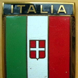 Può includere: Uno scudo in metallo dorato con la parola "ITALIA" in lettere nere in alto. Lo scudo è diviso in tre sezioni verticali: verde, bianco e rosso. La sezione bianca presenta una croce rossa su sfondo bianco.