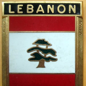May include: A rectangular pin featuring the flag of Lebanon. The top section reads "LEBANON" in gold letters on a black background. The flag design includes red stripes, a white center with a green cedar tree, and a gold border.