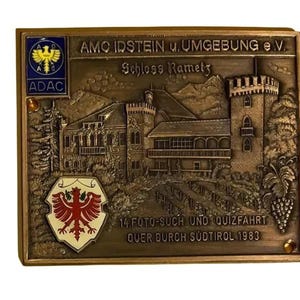 May include: A bronze-coloured rectangular plaque with embossed details. The plaque features a detailed depiction of a castle, German text, and two emblems. The text includes "AMC IDSTEIN u. Umgebung e.V." and "Schloss Rametz".