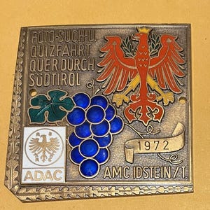 May include: Vintage metal plaque with text "FOTO-SUCH-U QUIZFAHRT QUER DURCH SÜDTIROL" and "1972". Features a red eagle, blue grapes, and the ADAC logo. The plaque has a gold-coloured border and is likely a souvenir or commemorative item.