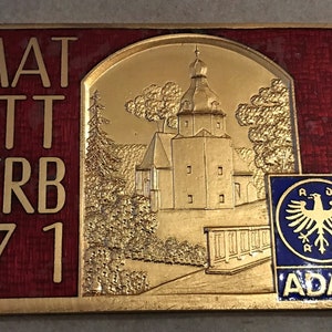 May include: A gold and red metal plaque with the text "HEIMAT WETT BEWERB 1971" and a gold image of a church with a spire. The plaque also has the text "ALTE KAPELLEN UND NEUE KIRCHEN" and a blue and gold logo with the text "ADAC" and a bird.