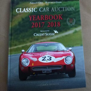 May include: A book titled "CLASSIC CAR AUCTION YEARBOOK 2017 2018" featuring a red vintage race car with the number 23. The book cover has text that reads "Sponsored by CREDIT SUISSE" and the authors' names.