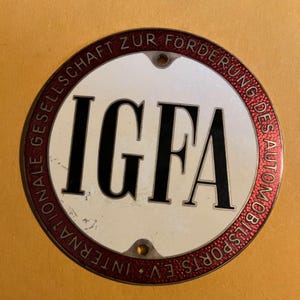 Puede incluir: Un emblema metálico redondo vintage con las letras "IGFA" en negrita negra. El emblema tiene un centro blanco, un borde rojo con texto y dos pequeños agujeros. El texto alrededor del borde dice "INTERNATIONALE GESELLSCHAFT ZUR FÖRDERUNG DES AUTOMOBILSPORTS E.V."