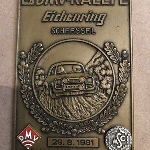 Puede incluir: Placa rectangular de color bronce con el texto "4. DMV-RALLYE Eichenring SCHEESSEL" en la parte superior. Un emblema circular central representa un coche de rally. La fecha "29.8.1981" está en la parte inferior. Hojas de roble y bellotas decoran la placa.
