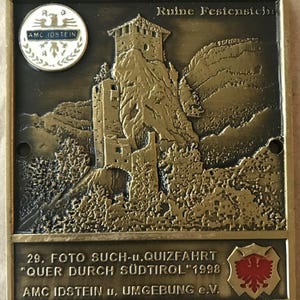 May include: A bronze-coloured metal plaque with a relief of a castle ruin on a rocky hillside. The plaque features German text, a circular emblem, and a shield with a red emblem. The text includes "Ruine Festenstein" and "29. FOTO SUCH-u. QUIZFAHRT 'QUER DURCH SÜDTIROL' 1998".