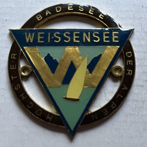 Könnte beinhalten: Eine goldfarbene Metallplakette mit einem blauen und grünen Dreieck in der Mitte. Das Dreieck hat einen goldenen Buchstaben "W" und ein gelbes Segelboot. Der äußere Kreis ist schwarz mit goldenen Buchstaben, die "BADESEE WEISSENSEE DER ALPEN HOCHSTER" lauten.