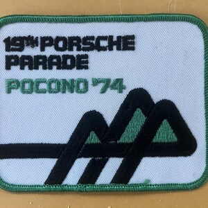 Könnte beinhalten: Rechteckiger Aufnäher mit weißem Hintergrund und grünem Rand. Der Text "19th PORSCHE PARADE POCONO '74" ist in Schwarz und Grün dargestellt. Eine schwarz-grüne Berggrafik befindet sich unten.