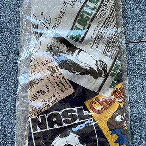 May include: A vintage tie featuring a collage of images related to soccer, including a soccer ball, a player's legs, and the text "NASL". The tie is wrapped in clear plastic.