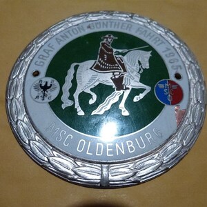 Könnte beinhalten: Eine silberne Metallplakette mit grünem Hintergrund, auf der ein Ritter zu Pferd abgebildet ist. Die Plakette hat einen silbernen Lorbeerkranz am Rand und den Text "Graf Anton Gunther Fahrt 1965 MSC Oldenburg" um die Mitte.