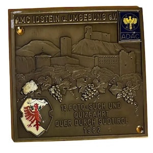 May include: A bronze plaque with embossed imagery of a castle, grape vines, and text. The plaque features the text "AMC IDSTEIN u. UMGEBUNG e.V." and "13. FOTO-SUCH UND QUIZFAHRT QUER DURCH SÜDTIROL 1982". An ADAC emblem is visible.