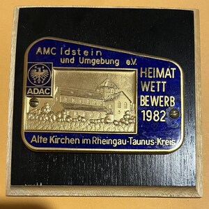 May include: A gold-coloured plaque with blue trim and a black background. The plaque features a church building, the text "AMC Idstein und Umgebung e.V.", "ADAC", "HEIMAT WETT BEWERB 1982", and "Alte Kirchen im Rheingau-Taunus-Kreis".