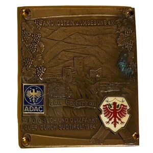 May include: A bronze-colored plaque with embossed details. The plaque features text, a castle illustration, and logos. Text includes "AMC IDSTEIN UMGEDUNG e.V.", "ADAC", "Burg Gacheppan", and "15. FOTO-SUCH UND QUIZFAHRT QUER DURCH SÜDTIROL 1984".