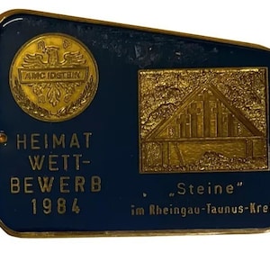 May include: A dark blue rectangular plaque with gold trim and text. It features a gold emblem with an eagle and the text "AMC IDSTEIN". Below, it reads "HEIMAT WETT-BEWERB 1984". A gold relief of a building is on the right, with the text "Steine".