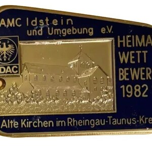 May include: A rectangular, gold-coloured metal pin with a dark blue border. The pin features text and a detailed image of a building. The text includes "AMC Idstein", "ADAC", "HEIMAT WETT BEWERB 1982", and "Alte Kirchen im Rheingau-Taunus-Kreis".