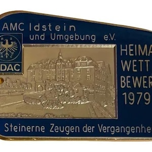 May include: A rectangular, gold-coloured metal pin with a blue border. The pin features a detailed relief of a building and text that reads "HEIMAT WETT BEWERB 1979". Additional text includes "AMC Idstein und Umgebung e.V." and "Steinerne Zeugen der Vergangenheit".