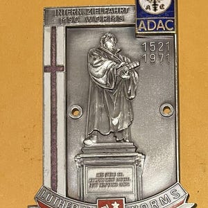 Puede incluir: Placa conmemorativa ADAC plateada que representa a Martín Lutero sosteniendo un libro. La placa incluye el texto "LUTHER IN WORMS" y "INTERN. ZIELFAHRT MSC WORMS". La esquina superior derecha muestra el logotipo de ADAC y los años 1521 y 1971.