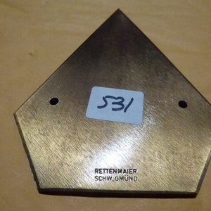 以下が含まれることがあります： 真鍮色の五角形のオブジェクトで、2つの穴と青いステッカーに「531」の数字があります。「RETTENMAIER SCHW.GMUND」の文字も表示されています。オブジェクトは、起毛した金属の質感を持っています。