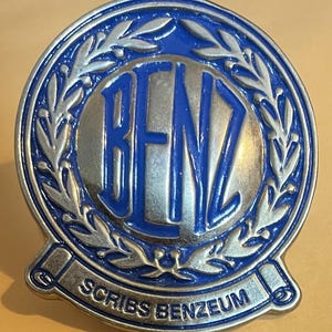 May include: A silver and blue metal emblem with a wreath of leaves surrounding a circle with the word "BENZ" in blue lettering. A silver banner below the circle reads "SCRIBS BENZEUM".