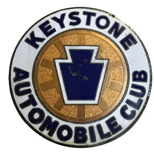 以下が含まれることがあります： キーストーン自動車クラブの丸いエンブレム。エンブレムは、青色の「KEYSTONE AUTOMOBILE CLUB」の文字が入った白い外側のリングが特徴です。内側には、中央に青いキーストーンの形をした金色のリングがあります。