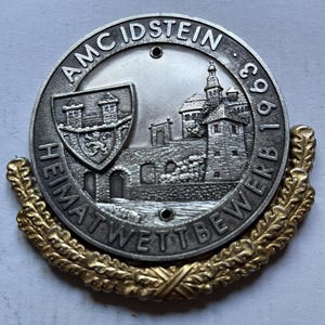 May include: A silver-coloured round medal with the text "AMC IDSTEIN 1963 HEIMATWETTBEWERB". The medal features a detailed relief of a castle and a coat of arms. A gold-coloured laurel wreath encircles the bottom half of the medal.