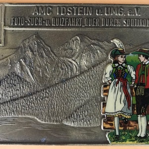 May include: A silver metal badge featuring a mountain scene and two people in traditional attire. The text on the badge reads "AMC IDSTEIN U. UMG. e.V. FOTO-SUCH-U. QUIZFAHRT QUER DURCH SODTIROL 1978."