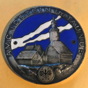 May include: A circular, silver-coloured metal emblem with a blue and white enamel background. It features a detailed illustration of a church and buildings, with the text "AMC IDSTEIN IM TAUNUS" around the edge. A smaller emblem with an eagle is at the bottom.