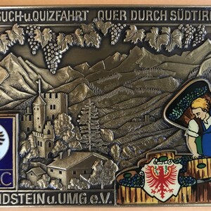 May include: A bronze plaque featuring a detailed image of a castle, mountains, and grapevines. The text "FOTO-SUCH-u.QUIZFAHRT QUER DURCH SÜDTIROL 1977" is at the top of the plaque. The text "ADAC" is at the bottom left of the plaque. The text "AMC IDSTEIN U.UMG.E.V." is at the bottom right of the plaque.