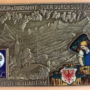 May include: A bronze plaque with a relief image of a vineyard landscape, a castle, and a person carrying grapes. The plaque has the text "FOTO-SUCH-u.QUIZFAHRT "QUER DURCH SÜDTIROL" 1977" at the top and "AMC IDSTEIN u.UMG.e.V." at the bottom. The plaque also features the ADAC logo.