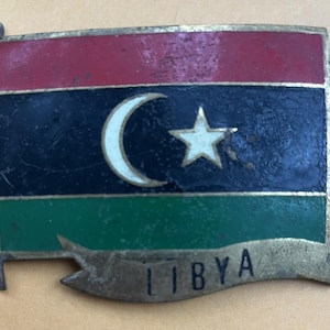 May include: A vintage metal pin depicting the flag of Libya. The flag design features red, black, and green stripes, with a white crescent and star in the centre. The word "LIBYA" is displayed on a banner below the flag.
