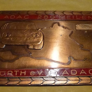 May include: A bronze-colored metal badge with a red border. The badge features a stylized image of a race car and a track map. The text "ADAC 280 MEILEN NÜRBURGRING HÜRTH e.V. für ADAC" is inscribed on the badge.