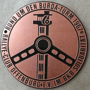 May include: A round, copper-colored metal emblem with black text and design elements. The emblem features a stylized tower design with the number "6" and the text "RUND UM DEN BURDA-TURM 1967" and "RALLYE-CLUB OFFENBURG E.V. IM RKB SOLIDARITAT".