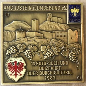 May include: A square, bronze-coloured metal plaque with embossed details. The plaque features a landscape with a castle, grape vines, and text that reads "AMC IDSTEIN u. UMGEBUNG e.V." and "13.FOTO-SUCH UND QUIZFAHRT QUER DURCH SÜDTIROL 1982". An ADAC logo is in the upper right corner.