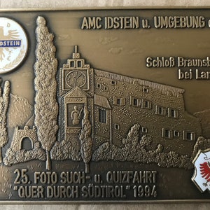 May include: A bronze plaque featuring an image of a castle and trees. The text on the plaque reads "AMC IDSTEIN u. UMGEBUNG e.V. Schloss Braunsberg bei Lana 25. FOTO SUCH- u. QUIZFAHRT "QUER DURCH SÜDTIROL" 1994".